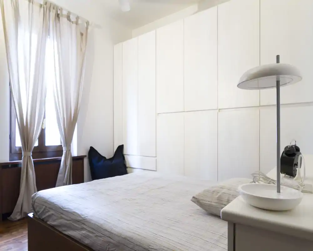 Camera da letto luminosa e minimal con letto matrimoniale ben rifatto, armadi a muro bianchi e luce naturale dalla finestra con tende.