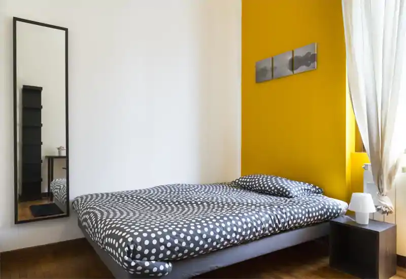 Dormitorio moderno y luminoso con cama doble con ropa de lunares, pared amarilla de acento, mesita con lámpara y espejo de cuerpo entero que refleja parte de la habitación.