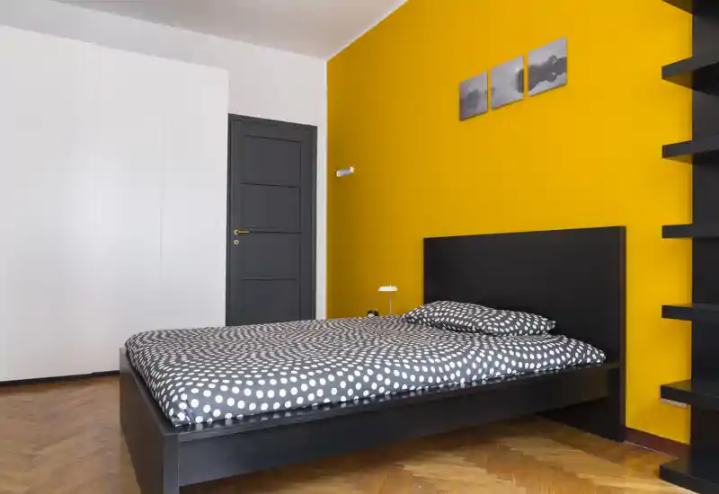 Camera da letto moderna e luminosa con letto basso nero, biancheria a pois e parete d'accento gialla. Spazio minimale, ben illuminato con armadio a muro e mensole sospese.
