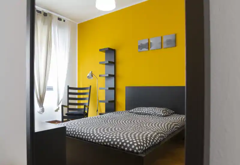 Camera da letto moderna e luminosa con letto matrimoniale, parete accent gialla e biancheria a pois; luce naturale da finestra e arredi neri minimalisti.