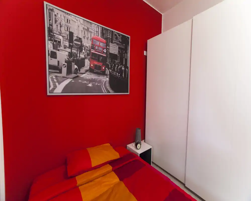 Camera da letto compatta con letto rosso e arancione, grande armadio bianco e comodino; arredamento luminoso e caldo con una stampa del bus di Londra sopra il letto.