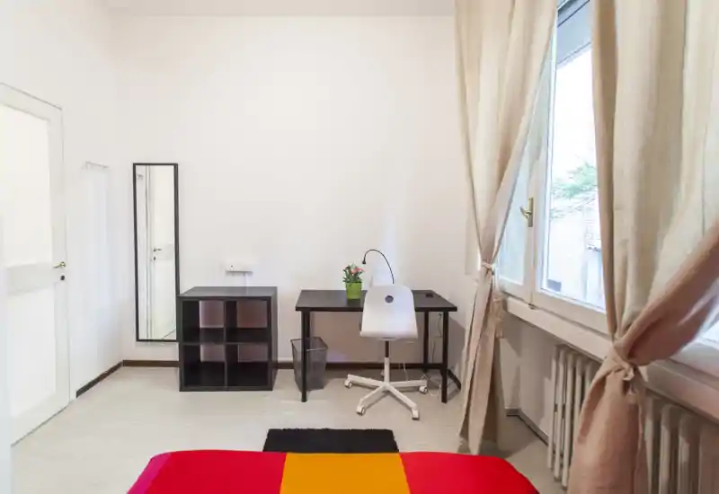 Camera da letto luminosa e minimal con copriletto colorato, una piccola scrivania e sedia vicino alla finestra e semplici contenitori. La luce naturale riempie la stanza rendendola ariosa e fresca.