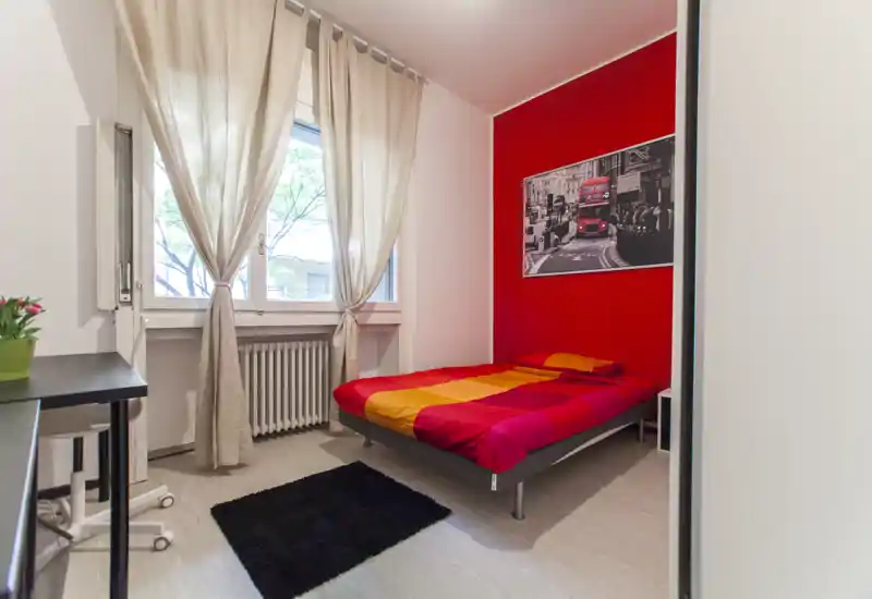 Camera singola accogliente con parete rossa a contrasto e biancheria colorata, ampia finestra con tende leggere, zona scrivania e piccolo tappeto — luminosa e ben arredata, ideale per un inquilino.