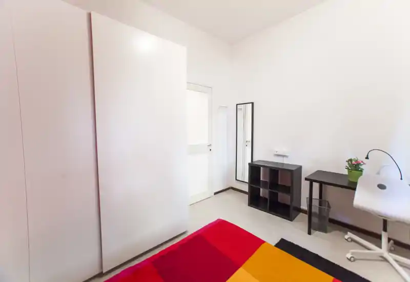 Camera da letto luminosa e ordinata con copriletto colorato, armadio, scrivania e specchio. Layout compatto e ben illuminato, ideale per una singola o uno studente.