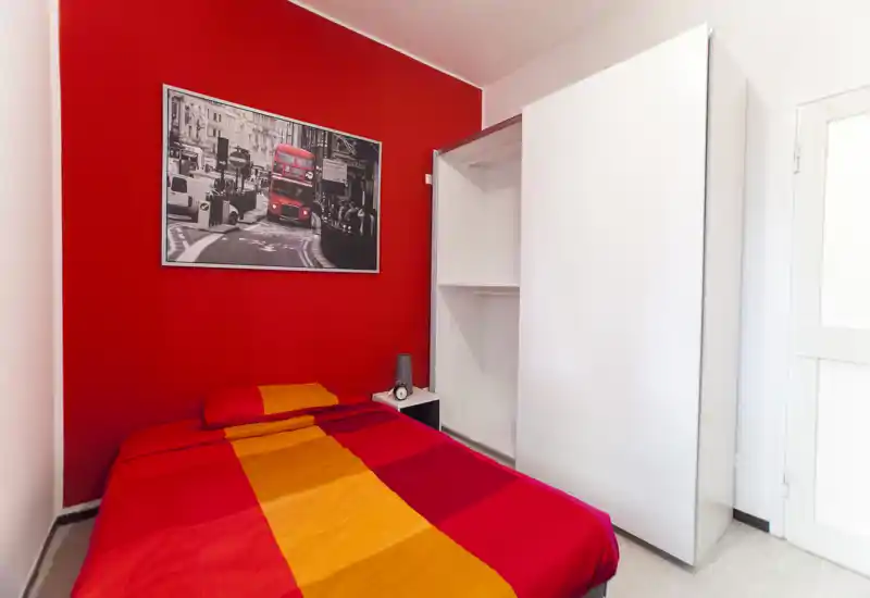 Camera da letto luminosa e vivace con letto matrimoniale dalle lenzuola rosse e arancioni su una parete d'accento rossa e un armadio bianco. L'ambiente è moderno e ben illuminato, con una stampa cittadina sopra il letto.