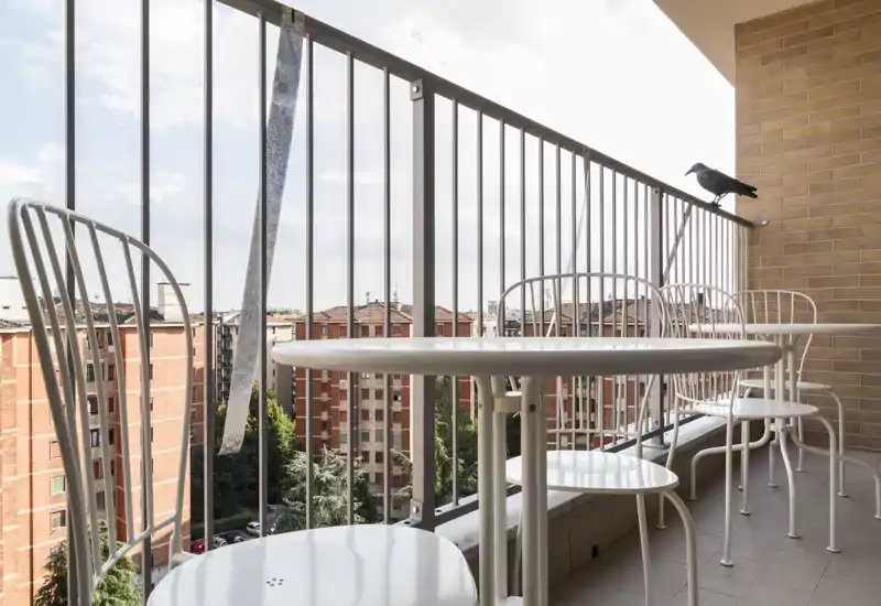 Balcón soleado con una pequeña mesa redonda y sillas metálicas a juego, con vistas a los edificios vecinos y las copas de los árboles — ideal para tomar un café por la mañana.