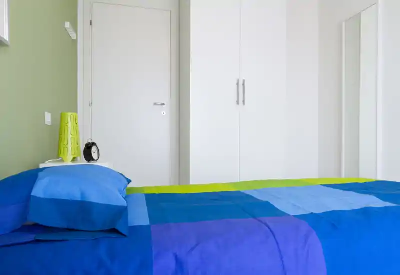 Dormitorio moderno y luminoso con colcha azul y verde, mesita con lámpara y despertador y armario blanco — ordenado y bien iluminado para un anuncio.