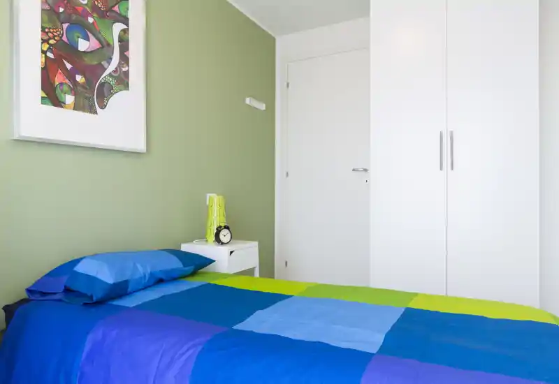 Dormitorio individual luminoso y moderno con edredón azul y verde, mesita con lámpara y reloj, cuadro enmarcado y armario blanco: limpio, bien iluminado y acogedor.