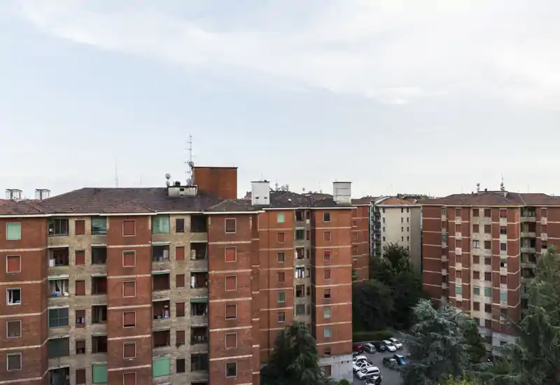 Vista exterior de edificios residenciales de ladrillo con balcones y un pequeño aparcamiento, tomada con luz suave: adecuada para mostrar la fachada y el entorno del barrio.