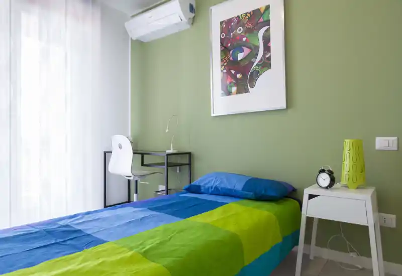 Habitación individual luminosa y ordenada con ropa de cama azul y verde, mesita con lámpara y reloj, un pequeño escritorio con silla y aire acondicionado en la pared.