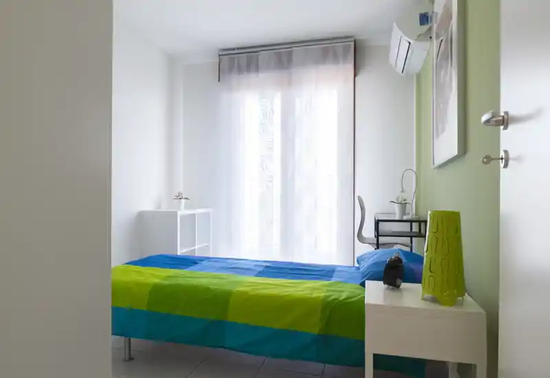 Dormitorio luminoso y compacto con cama vestida en tonos azul y verde, mesita de noche, pequeño escritorio y una gran ventana con cortina translúcida que baña la estancia de luz natural.