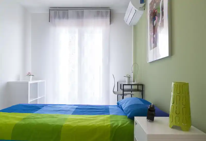 Dormitorio moderno y luminoso con cama colorida, mesita y un pequeño escritorio junto a una ventana con cortina translúcida; ambiente ordenado y agradable.