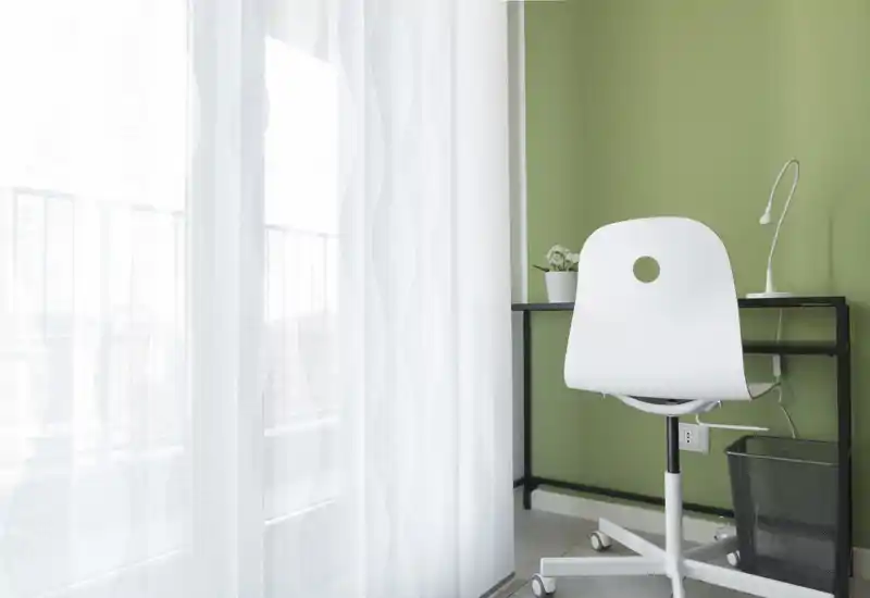 Rincón de trabajo luminoso en el salón con una silla giratoria blanca, escritorio negro compacto, lámpara de escritorio y una planta pequeña junto a una gran ventana con cortina translúcida que deja pasar luz natural.