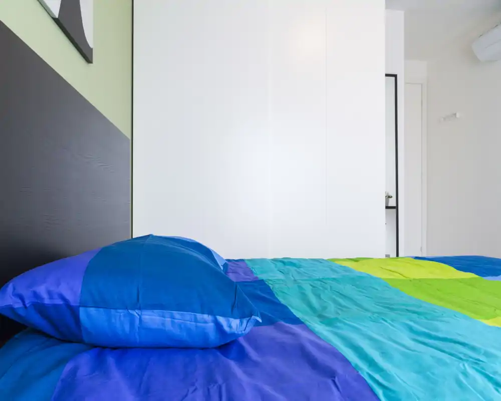 Camera da letto moderna e luminosa con biancheria colorata e un ampio letto che occupa gran parte dell'inquadratura; armadio bianco pulito e arredo minimalista per un ambiente fresco e accogliente.