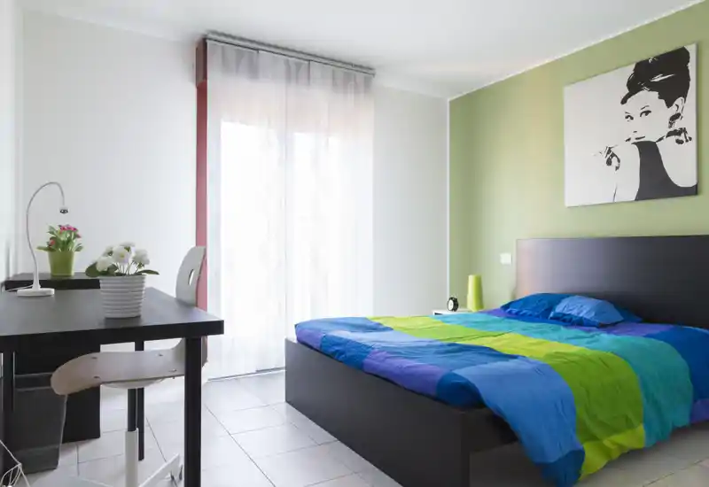 Camera da letto moderna e luminosa con un ampio letto dal copriletto colorato, lampada da comodino e scrivania vicino a una finestra con tenda leggera — perfetta per un soggiorno confortevole.