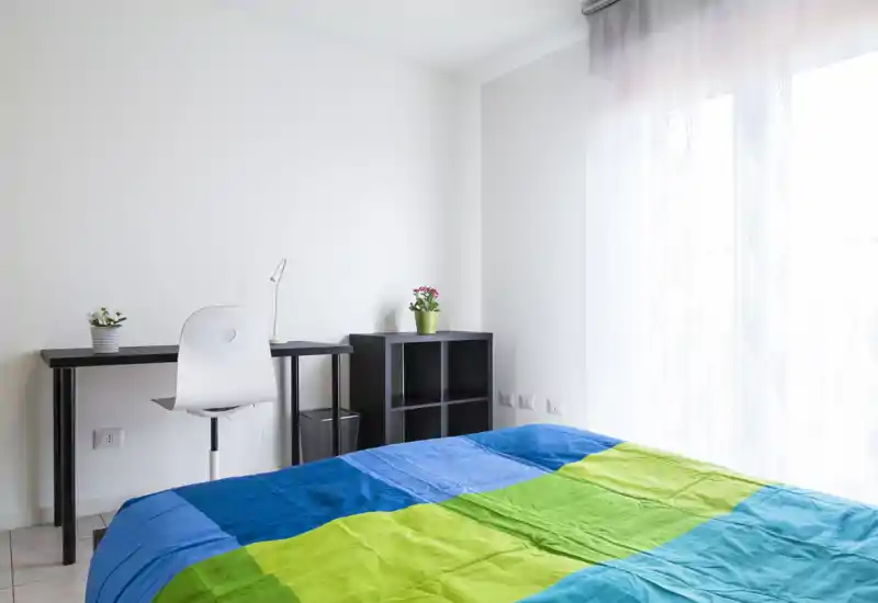 Camera da letto luminosa e minimal con una coperta colorata, una scrivania e una sedia vicino a una grande finestra che illumina l'ambiente con luce naturale.
