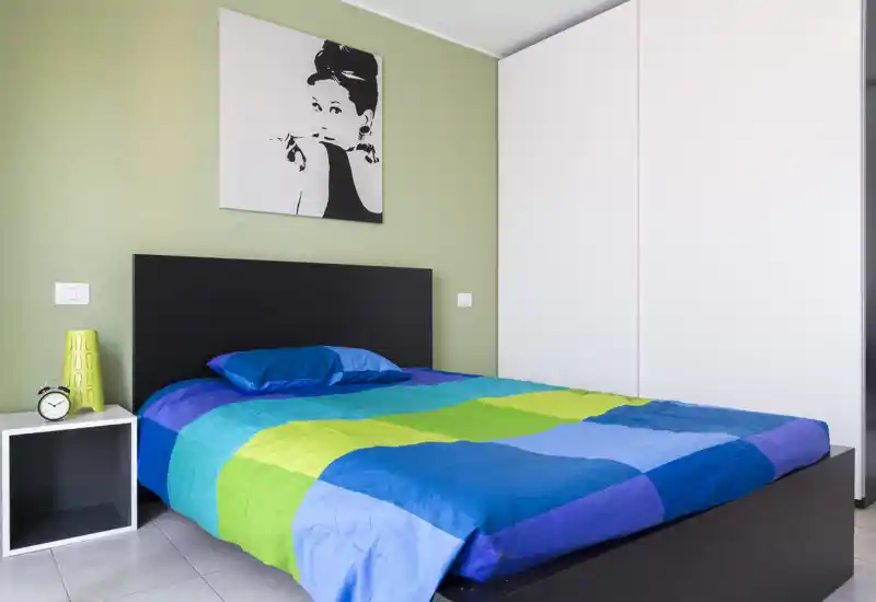 Camera da letto moderna e luminosa con un letto matrimoniale colorato, testiera nera e grande armadio bianco. Linee pulite, luce naturale e arredo minimale creano un ambiente fresco e accogliente, perfetto per annunci.