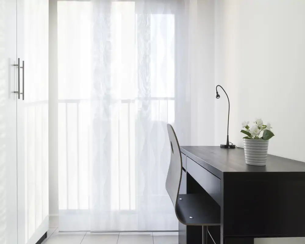 Espace de vie lumineux et minimaliste avec portes-fenêtres voilées, un petit bureau noir avec chaise, une lampe de bureau et une plante en pot pour une touche de verdure.
