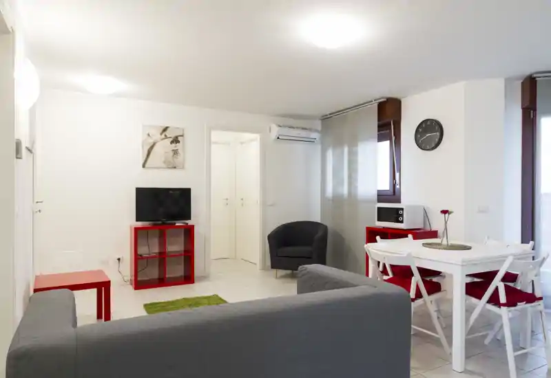 Zona de estar moderna y luminosa con sofá gris, butaca, televisión sobre mueble rojo y mesa de comedor blanca con cojines rojos — espacio ordenado y bien iluminado, ideal para grupos pequeños.