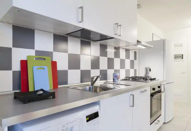 Cocina moderna y luminosa con fregadero de acero, placa de gas, horno integrado y mobiliario blanco; tablas de cortar coloridas aportan un toque de color sobre el salpicadero a cuadros.
