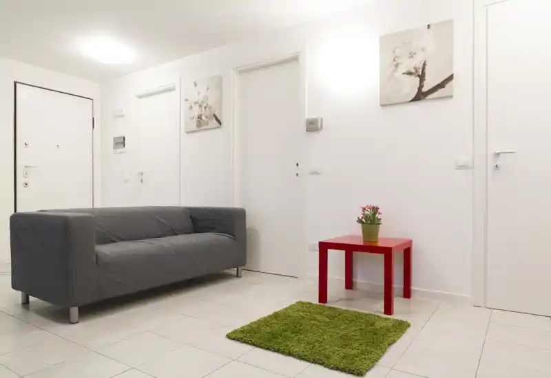 Entrada luminosa y minimalista con un sofá gris moderno, puertas blancas y una pequeña mesa roja con una planta — suelo de baldosas limpio y arte de pared sencillo.