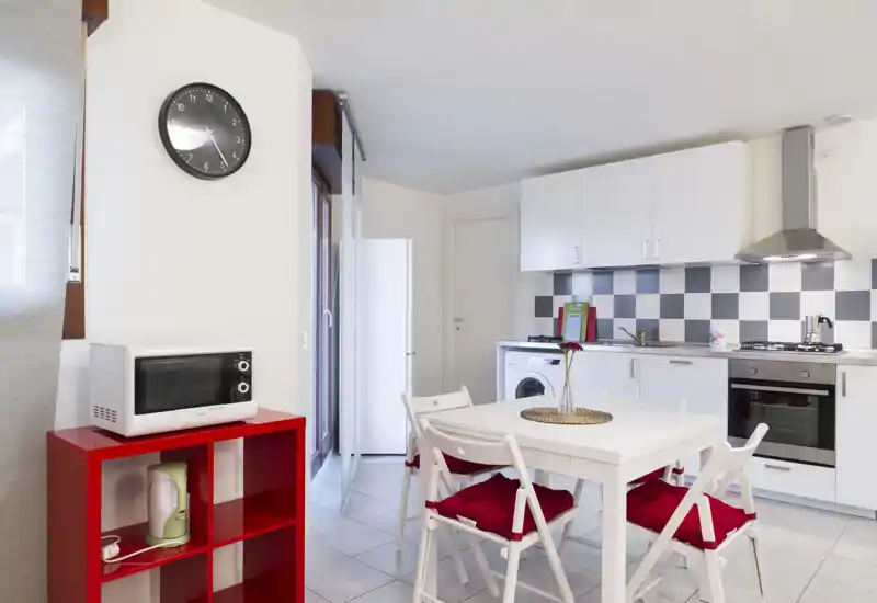 Cocina moderna y luminosa con muebles blancos, salpicadero a cuadros y una mesa pequeña con sillas con cojines rojos — distribución funcional con microondas, nevera y horno integrado.