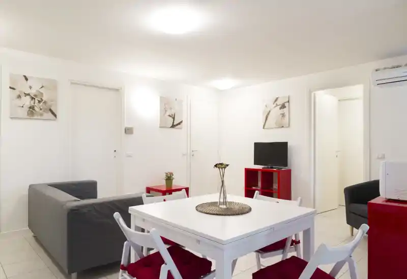 Área de estar moderna y luminosa con mesa blanca, cuatro sillas con cojines rojos, sofá gris y pequeño mueble de TV — estilo minimalista y buena iluminación.