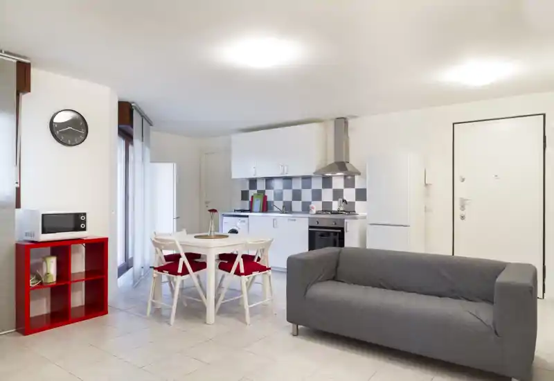 Zona de estar de planta abierta, luminosa y moderna con cocina integrada, mesa de comedor y un cómodo sofá gris; detalles rojos que aportan un aspecto fresco y acogedor.