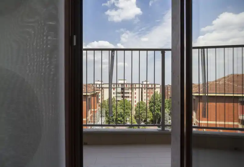 Small balcony with metal railing overlooking neighboring apartment buildings and treetops, seen through an open sliding door under a partly cloudy sky.