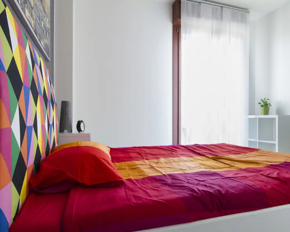 Chambre moderne et lumineuse avec une tête de lit géométrique colorée et un grand lit double habillé de linge rouge et orange. La lumière naturelle traverse un voile translucide, créant une ambiance fraîche et accueillante.