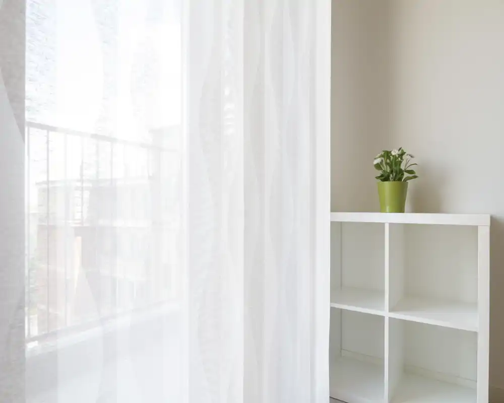 Un voile léger diffuse la lumière d'un petit balcon ; la balustrade et des bâtiments voisins sont visibles à travers le tissu. À droite, une étagère blanche à cases avec une petite plante apporte une touche aérienne et calme.