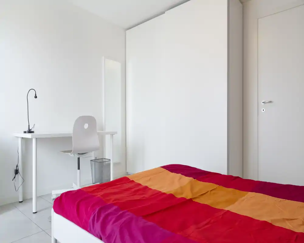 Chambre lumineuse et minimaliste avec une couette colorée, une armoire blanche et un petit bureau — design simple et épuré, idéal pour une personne.