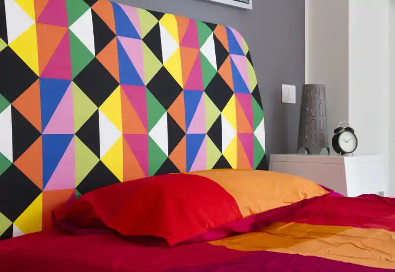 Primer plano de dormitorio colorido con cabecero geométrico, ropa de cama roja y naranja y mesita con lámpara y despertador; composición luminosa y alegre, ideal para un anuncio.