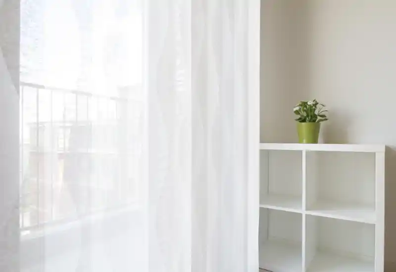 Una cortina translúcida difumina la luz de un pequeño balcón; se aprecia la barandilla y edificios cercanos a través de la tela. A la derecha, una estantería blanca con una planta crea un rincón sereno y luminoso.