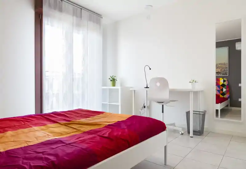 Dormitorio moderno y luminoso con edredón rojo-naranja llamativo, estructura de cama blanca y un escritorio junto a la ventana con luz natural.