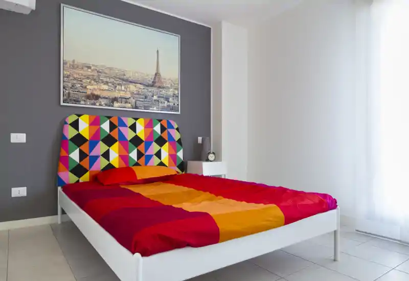 Dormitorio moderno y luminoso con cabecero geométrico colorido y ropa de cama roja-naranja vibrante. Gran ventana que aporta abundante luz natural; el espacio se ve cuidado y fresco.