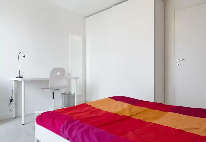 Dormitorio luminoso y minimalista con una colcha colorida, armario blanco y un pequeño escritorio: diseño sencillo y limpio, ideal para una persona.