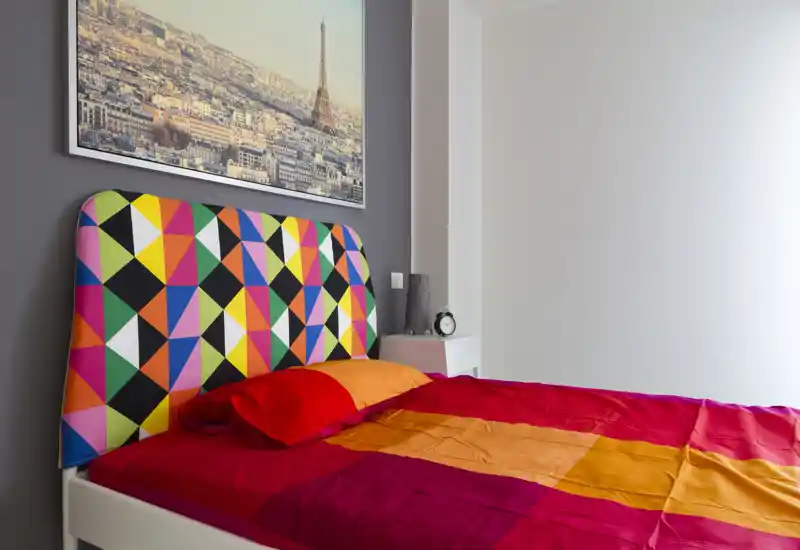 Dormitorio luminoso y colorido con cabecero tapizado geométrico y ropa de cama rojo-anaranjada vibrante; cama ordenada ideal para un alquiler con estilo.