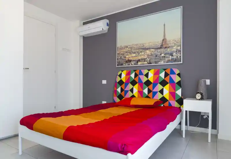 Dormitorio moderno y colorido con cama completa, cabecero geométrico vibrante, mesita con lámpara y reloj, y cuadro de la ciudad encima. Espacio luminoso y perfecto para alquileres de corta estancia.