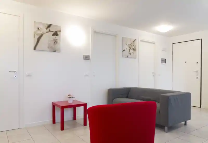 Bright, minimal living area with a grey sofa, red armchair and small red side table; neutral white walls and simple wall art create a clean, contemporary feel.