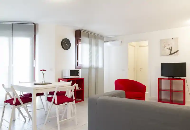 Bright, modern living area with a grey sofa, red accent armchair and a small dining table; clean minimalist styling with cheerful red details.