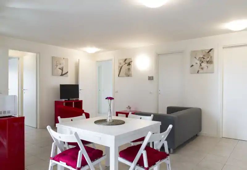 Bright, modern living area with a white dining table, four chairs with red cushions, a gray sofa and tasteful wall art — ideal for a compact apartment.