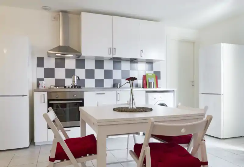 Bright modern kitchen with a white cabinetry and a small dining table with red-cushioned chairs. The space features stainless-steel appliances, a checkerboard backsplash and a tidy counter with a decorative vase.