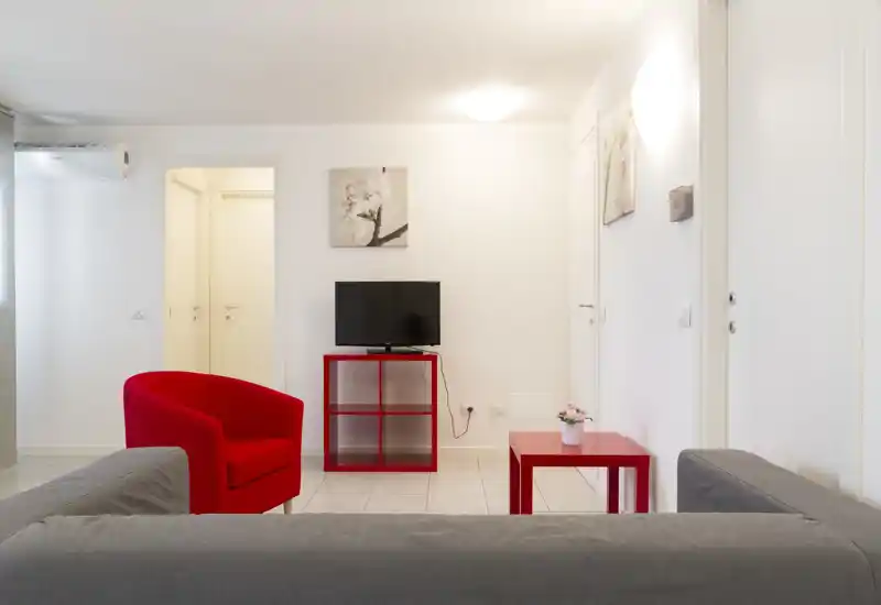 Bright, modern living area with a grey sofa, a red armchair and matching red tables centered around a small TV — clean minimalist styling and pleasant natural light.
