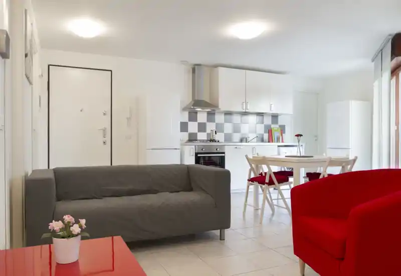 Bright, modern open-plan living area with a grey sofa, red armchair and an integrated white kitchen and dining table — ideal for a compact apartment.