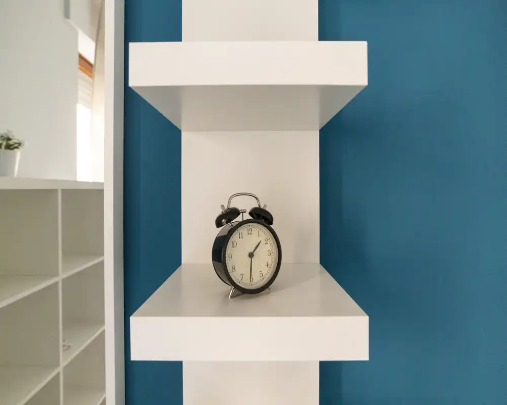 Close-up of a white wall shelf with a vintage black alarm clock against a blue wall; a shelving unit is partially visible at left. The photo highlights a decorative accessory and shelf details rather than a specific room.