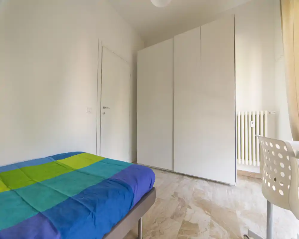 Bright, minimal bedroom with a double bed dressed in colorful bedding, a large white wardrobe and a work chair by the window — simple and airy.