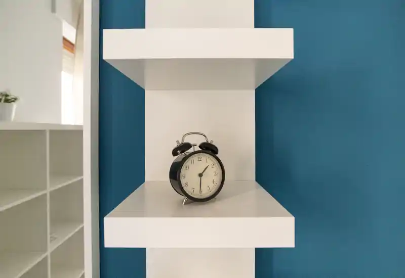 Close-up of a white wall shelf with a vintage black alarm clock against a blue wall; a shelving unit is partially visible at left. The photo highlights a decorative accessory and shelf details rather than a specific room.