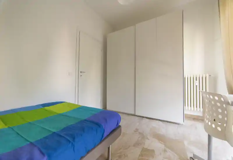Bright, minimal bedroom with a double bed dressed in colorful bedding, a large white wardrobe and a work chair by the window — simple and airy.