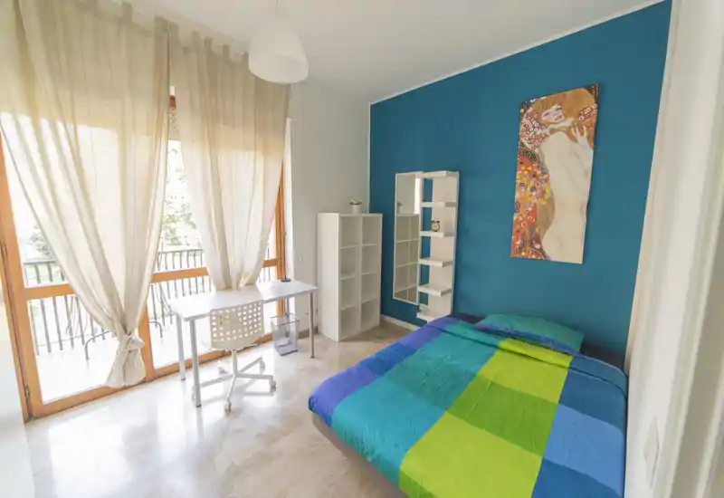 Bright, colorful bedroom with a double bed against a teal accent wall, shelving unit and a small desk by large windows letting in abundant natural light.